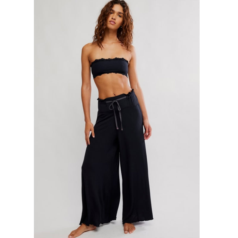 A woman modeling black wide-leg pants with a drawstring waist and a black strapless bandeau top against a light gray studio background.
