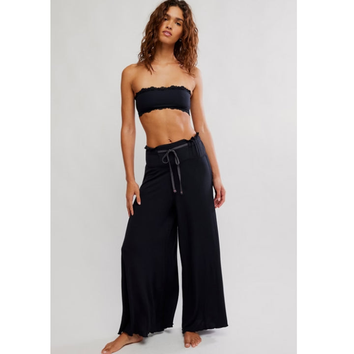 A woman modeling black wide-leg pants with a drawstring waist and a black strapless bandeau top against a light gray studio background.