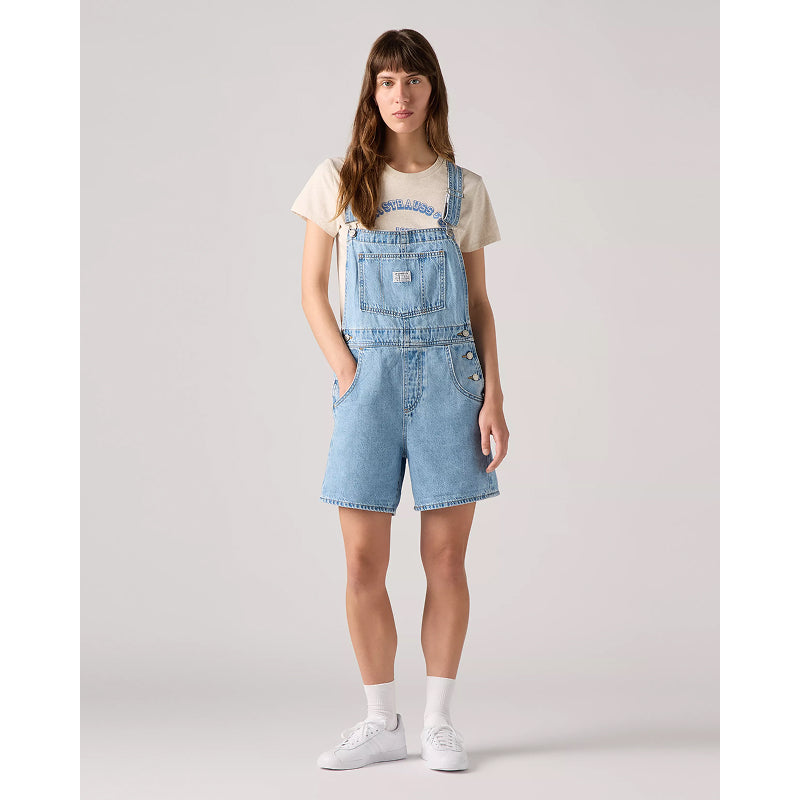 Woman wearing a light blue denim overall dress on a plain background