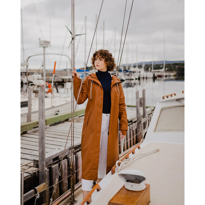 A woman standing by a sailboat wearing a brown raincoat with a V-neck sweater and white pants.